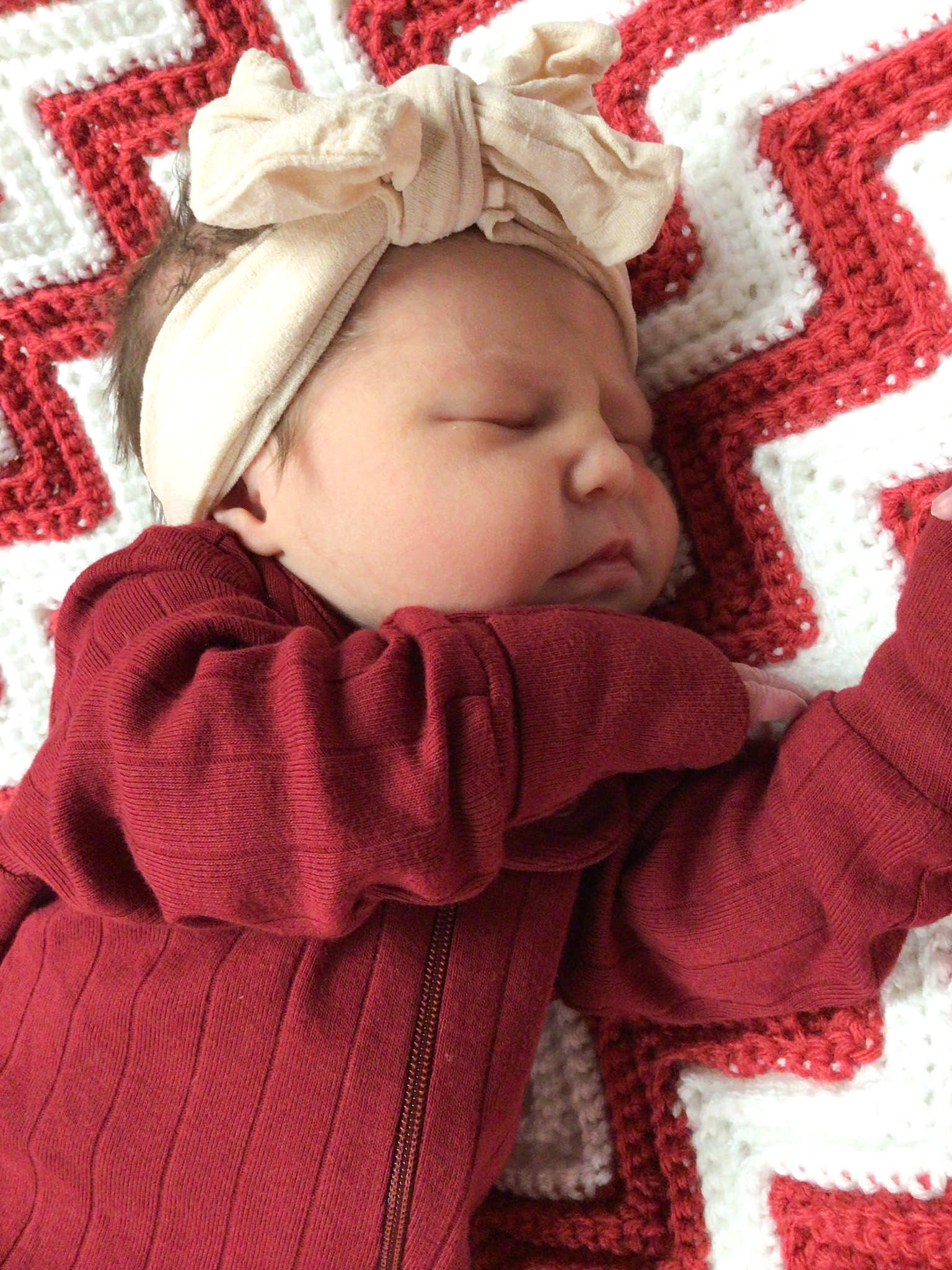 Sleeping baby in red outfit with cream bow headband on a red and white crochet blanket.