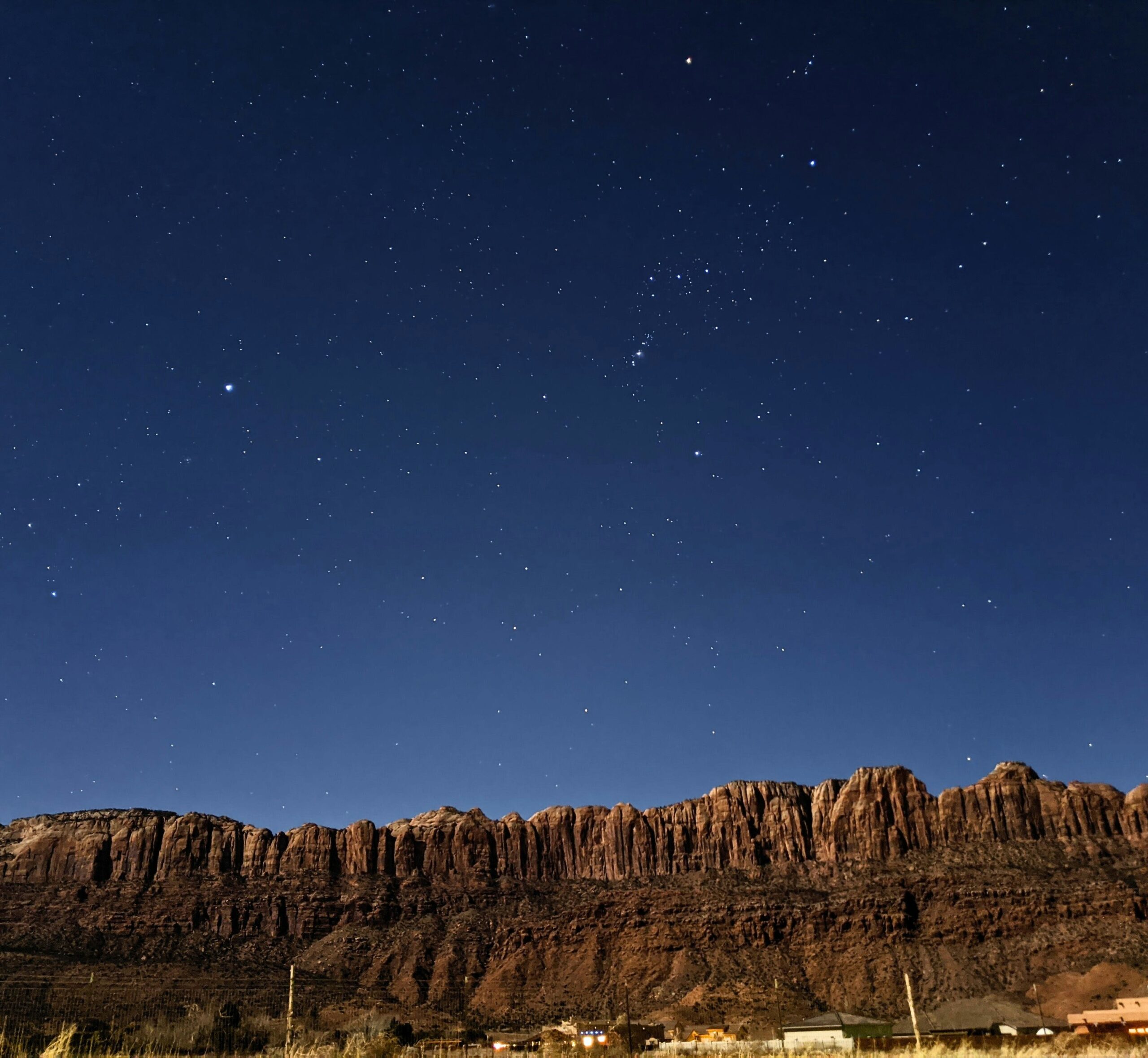Starry night sky over a rugged mountain ridge with a few lights below, showcasing natural beauty and tranquility.