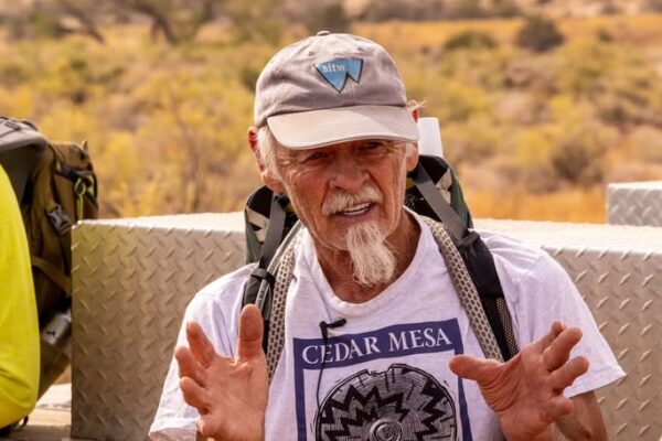 Joe Pachak in cap and backpack talking outdoors, wearing Cedar Mesa shirt in a desert landscape.