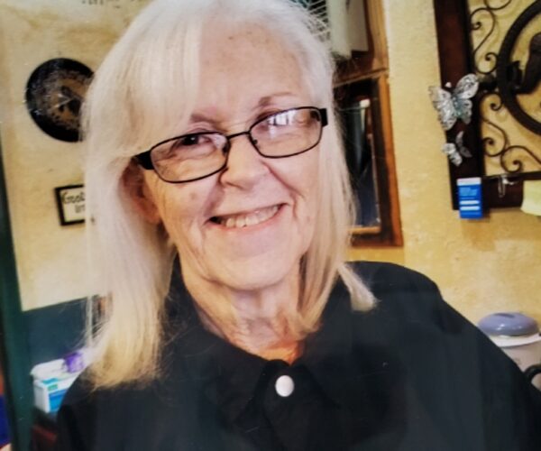 Smiling elderly woman with glasses and long hair in a cozy room setting, wearing a dark outfit.