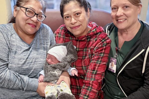 Family and nurse smiling with newborn baby wrapped in cozy outfit inside hospital room.