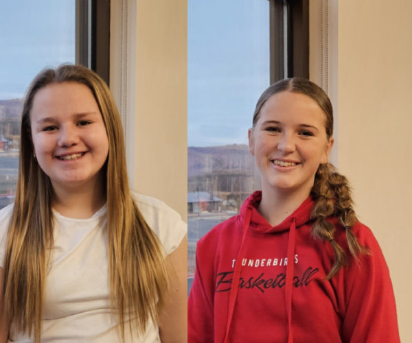 Two smiling girls indoors, one in a white shirt and the other in a red hoodie, with an American flag outside the window.