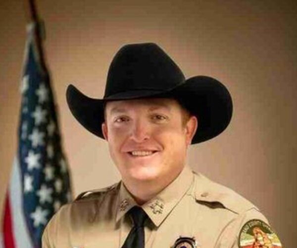 jamison wiggins in sheriff uniform and cowboy hat stands smiling with American flag in background.