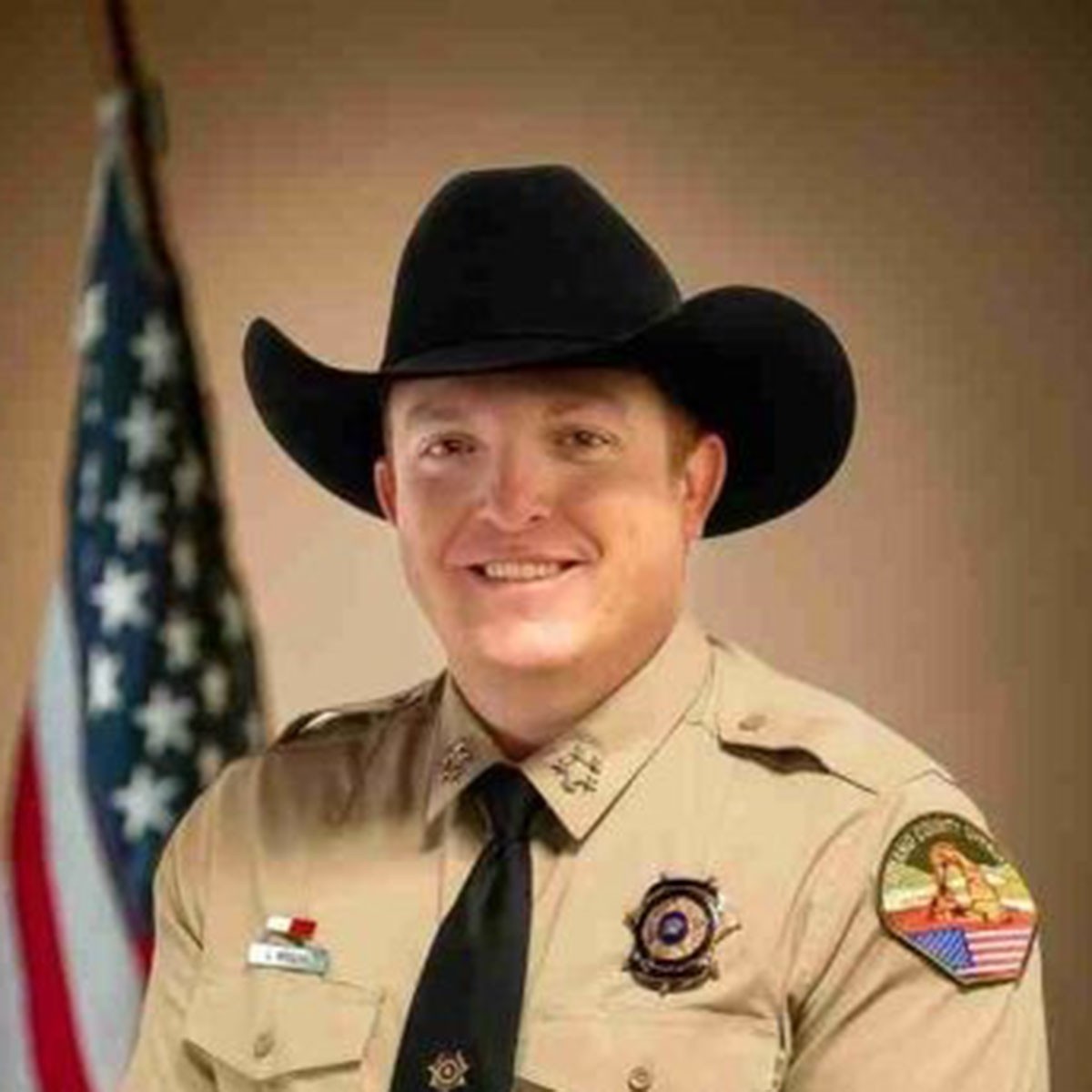 jamison wiggins in sheriff uniform and cowboy hat stands smiling with American flag in background.