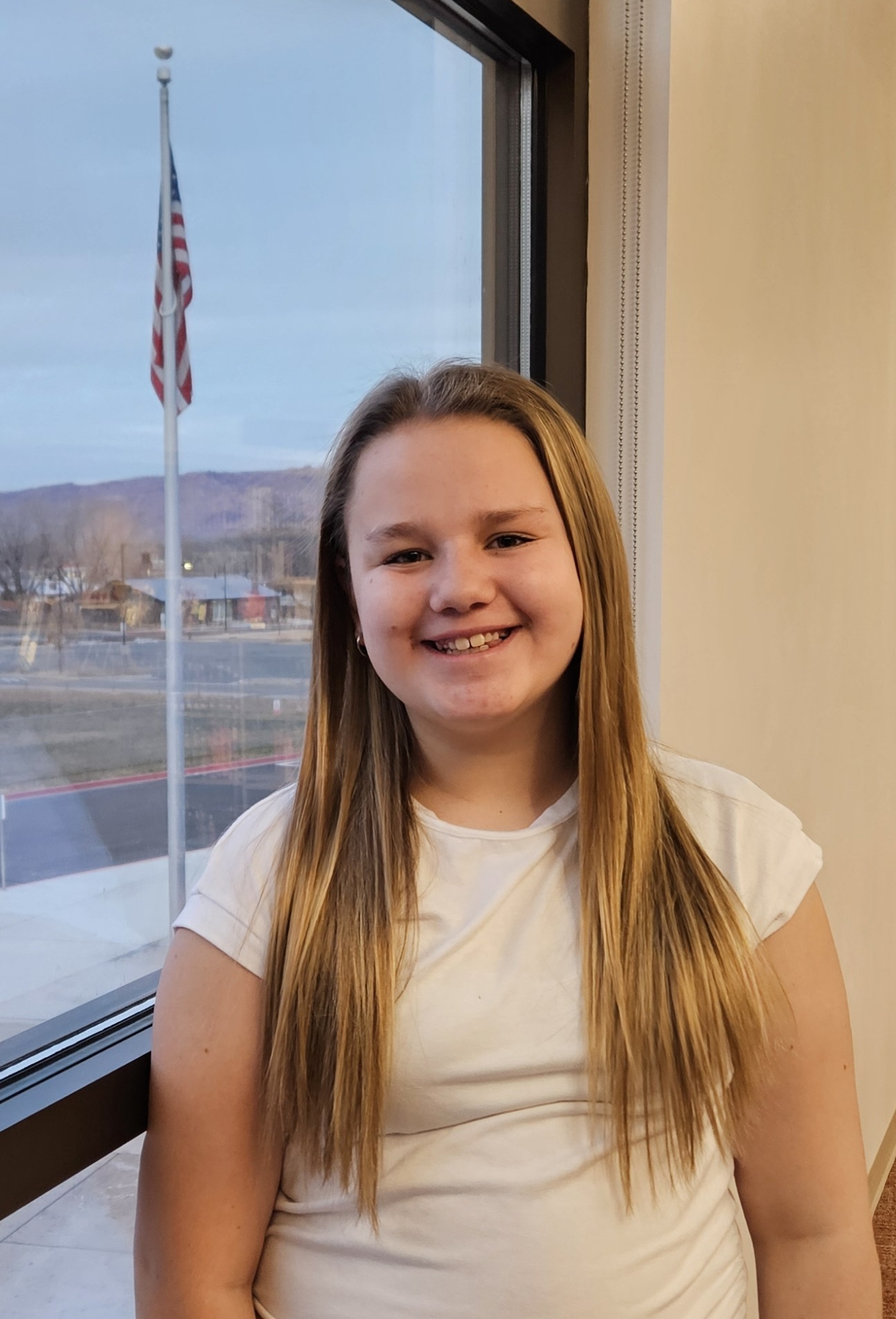 Smiling girl in a white shirt by a window, with an American flag and scenic view in the background.