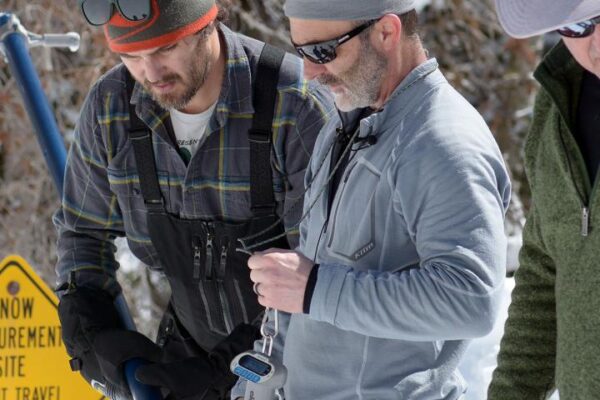 Three men measuring snow depth in winter, wearing outdoor gear near a yellow warning sign.